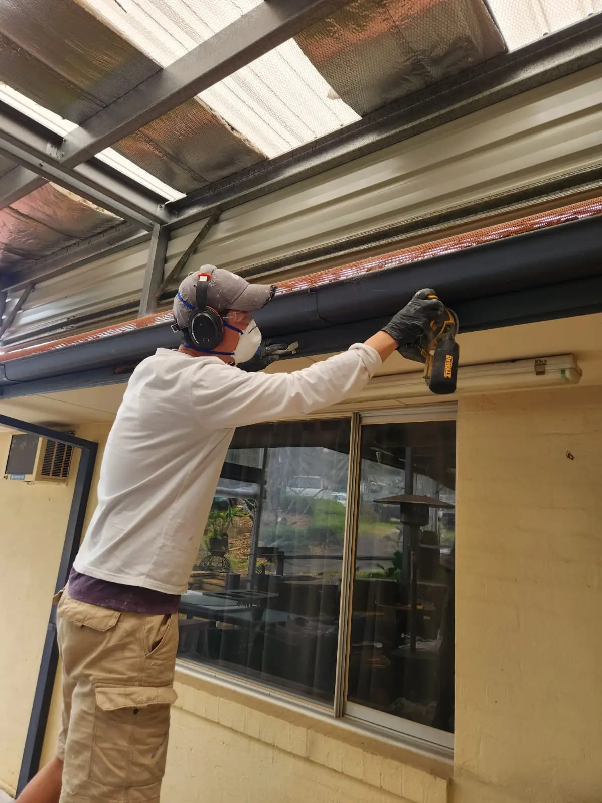 Painter in a respirator and ear muffs preparing metal guttering under a pergola